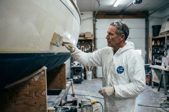 Artisan appliquant du gel-coat sur une coque en fibre de verre avec un pinceau large dans un atelier de réparation nautique