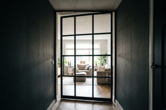 Porte verrière battante en acier noir installée au bout d'un couloir sombre avec vue sur un salon lumineux