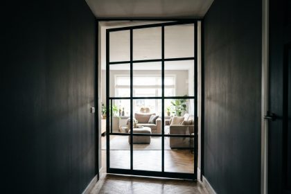 Porte verrière battante en acier noir installée au bout d'un couloir sombre avec vue sur un salon lumineux